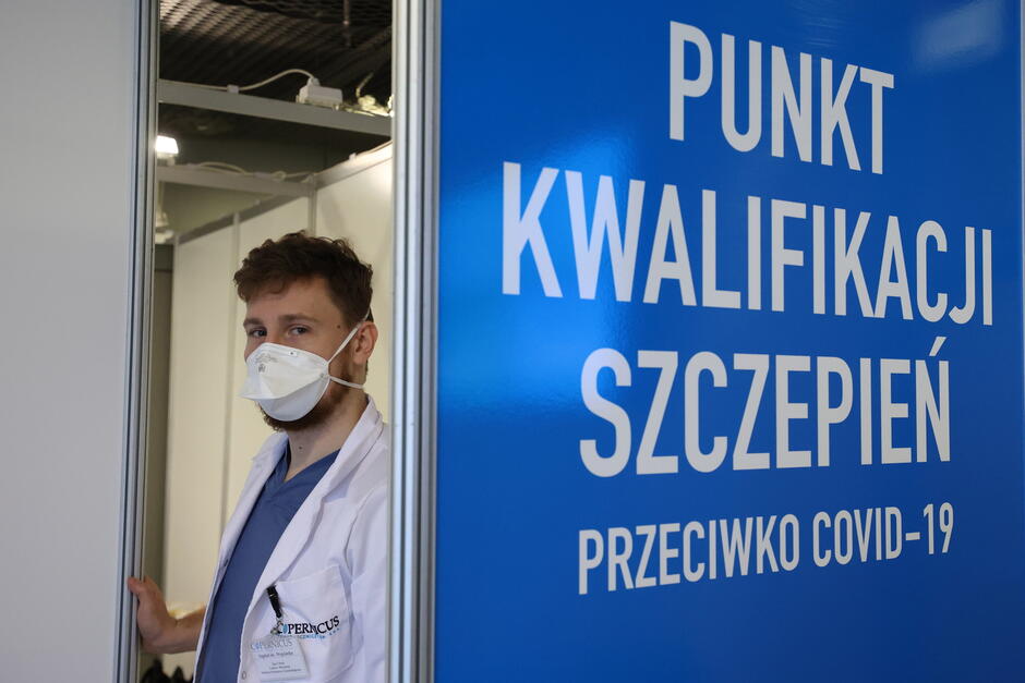 Zdjęcie przedstawia moment podawania szczepionki, gdzie ręce osoby wykonującej zastrzyk są ubrane w niebieskie rękawiczki ochronne. Igła wstrzykiwana jest w ramię osoby ubranej w czerwoną koszulkę, symbolizując procedurę medyczną w bezpiecznych warunkach.