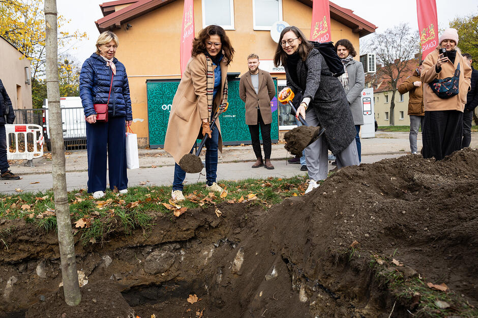 Na zdjęciu widzimy grupę ludzi uczestniczących w uroczystości sadzenia drzewa, gdzie dwie kobiety na pierwszym planie trzymają łopaty i symbolicznie zakopują ziemię. W tle widać inne osoby, które obserwują wydarzenie i robią zdjęcia, a cała scena odbywa się w miejskiej scenerii z budynkami i drzewami.