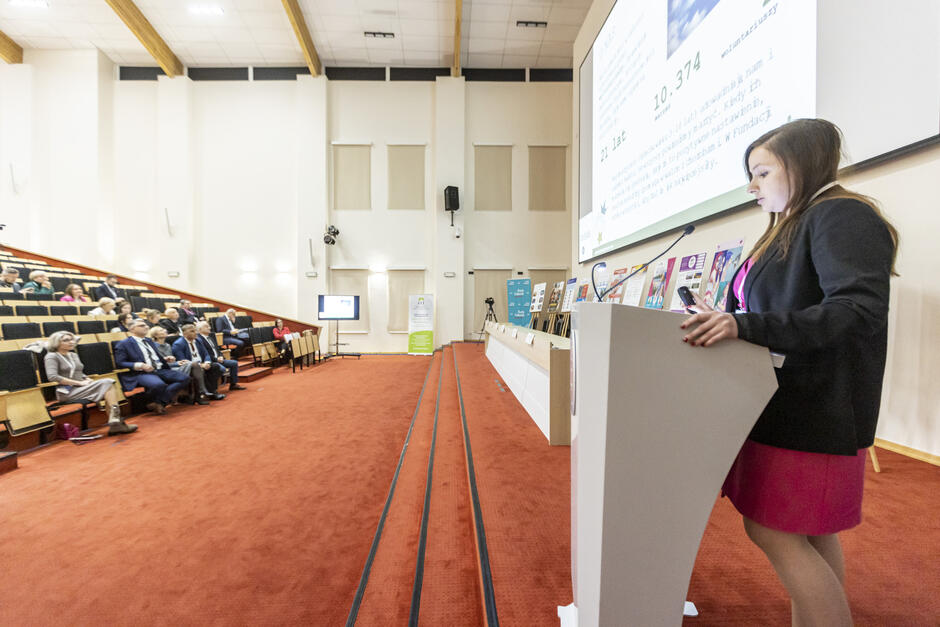 Na zdjęciu widać prelegentkę stojącą przy mównicy w dużej sali konferencyjnej, prezentującą treści na ekranie projekcyjnym. Przed nią siedzi publiczność, która uważnie słucha prezentacji, a sala utrzymana jest w oficjalnym, profesjonalnym stylu.