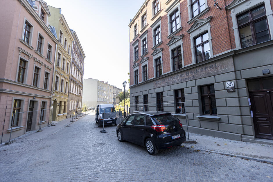 na zdjęciu dwie kamienice stojące po obu stronach ulicy, po prawej stoi samochód osobowy