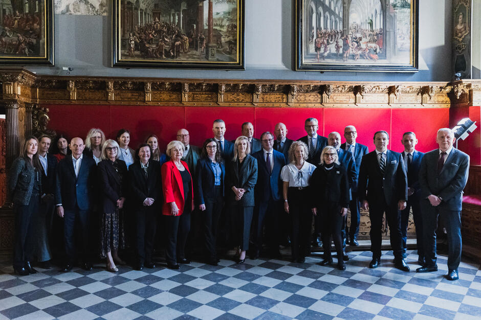 Na zdjęciu widoczna jest grupa osób ubranych formalnie, pozujących do wspólnego zdjęcia na tle ozdobnej, czerwonej ściany oraz zabytkowych malowideł. Sceneria sugeruje, że fotografia została wykonana w eleganckim, historycznym wnętrzu, co podkreśla oficjalny charakter spotkania.