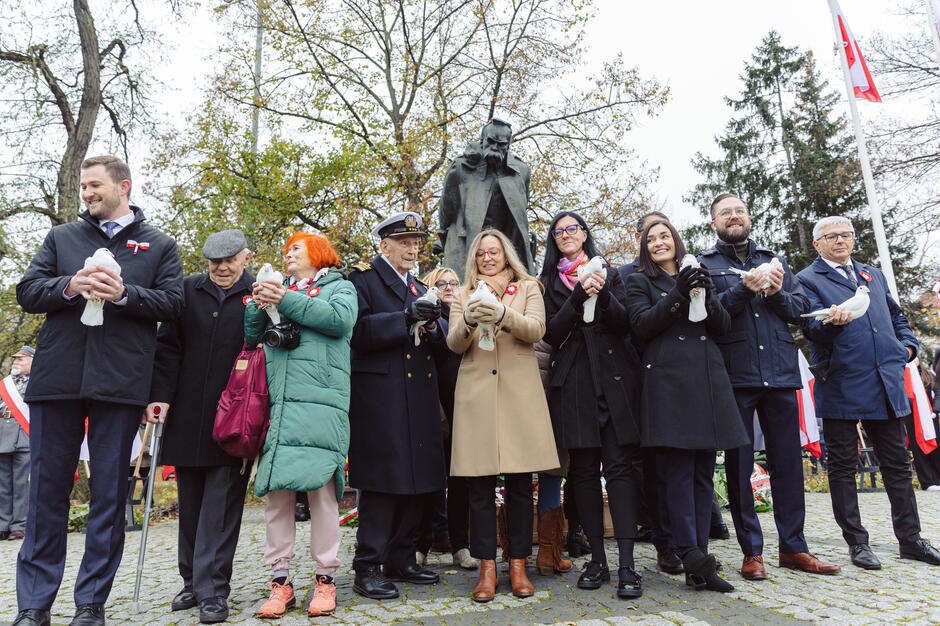 Na zdjęciu grupa osób stoi w rzędzie, trzymając białe gołębie, co symbolizuje pokój i może być częścią ceremonii lub uroczystości patriotycznej. W tle widać pomnik oraz flagi, a uczestnicy mają przypięte biało-czerwone kokardki, co sugeruje wydarzenie upamiętniające ważną dla Polski okazję