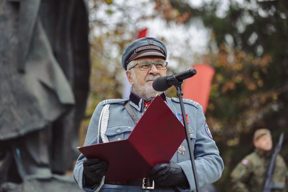 Na zdjęciu starszy mężczyzna w mundurze wojskowym przemawia do mikrofonu, trzymając czerwony folder. Ubrany w historyczny strój, z odznaczeniami na piersi, wygląda na osobę odgrywającą ważną rolę podczas patriotycznej ceremonii.