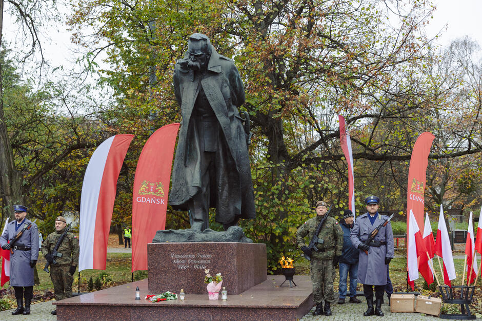 Na zdjęciu widzimy pomnik Józefa Piłsudskiego otoczony flagami Polski i Gdańska, ustawiony podczas uroczystości patriotycznej. Po obu stronach pomnika stoją żołnierze oraz strażnicy w historycznych mundurach, symbolizując hołd składany dla marszałka.