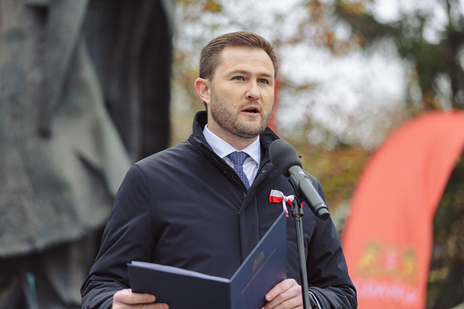 Na zdjęciu widzimy mężczyznę przemawiającego do mikrofonu podczas wydarzenia na świeżym powietrzu. Ubrany formalnie, z biało-czerwoną wstążką przypiętą do ubrania, co może sugerować, że bierze udział w patriotycznym lub państwowym wydarzeniu.






