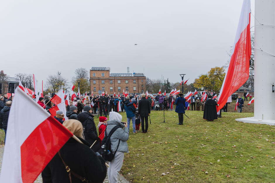 Na zdjęciu widać tłum uczestników zgromadzonych podczas uroczystości patriotycznej z okazji podniesienia polskiej flagi. Ludzie trzymają biało-czerwone flagi, a na pierwszym planie widać wielką flagę Polski przygotowaną do podniesienia na maszcie