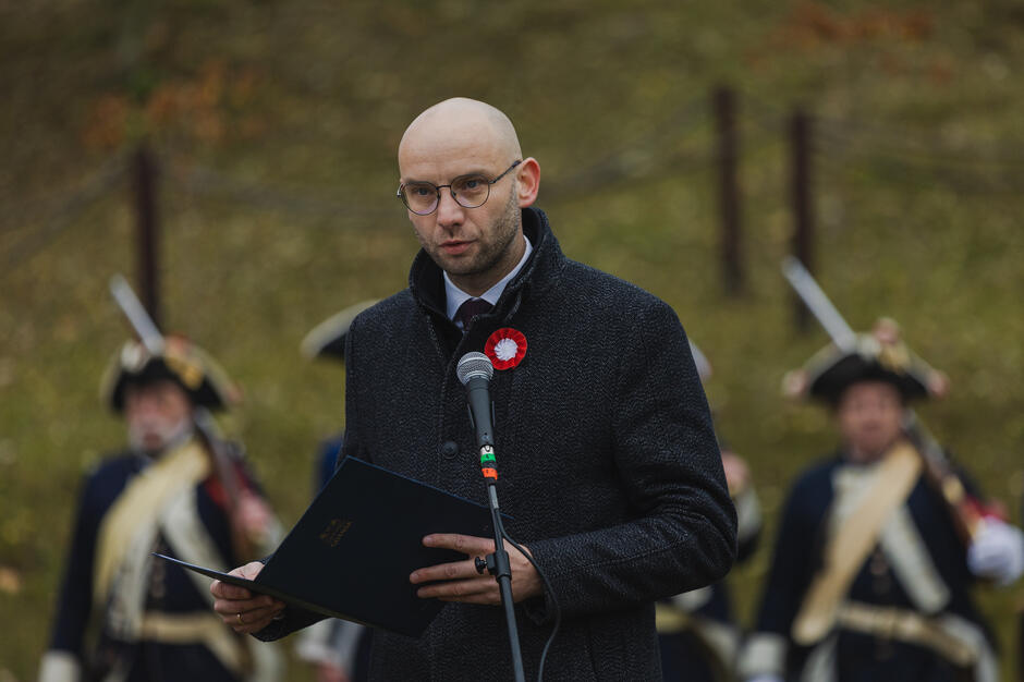 Na zdjęciu znajduje się mężczyzna przemawiający podczas uroczystości, ubrany w ciemny płaszcz i ozdobiony biało-czerwoną kokardą. W tle widać rekonstruktorów w historycznych strojach, co nadaje wydarzeniu patriotycznego i tradycyjnego charakteru.