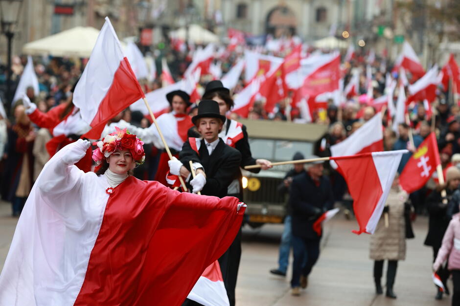 Na zdjęciu widoczna jest grupa uczestników parady ubrana w stroje stylizowane na biało-czerwoną flagę Polski, z wieloma osobami trzymającymi flagi narodowe. Uśmiechnięta kobieta na pierwszym planie ma na głowie wianek z kwiatów, co podkreśla radosny i uroczysty nastrój obchodów Święta Niepodległości.