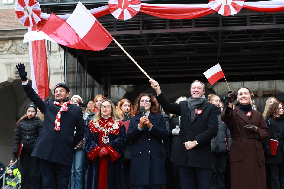 Na zdjęciu widoczna jest grupa osób stojących na scenie, uśmiechniętych i machających biało-czerwonymi flagami, w otoczeniu patriotycznych dekoracji. Wśród uczestników znajdują się osoby w oficjalnych strojach oraz miejskich insygniach, co wskazuje na udział przedstawicieli władz podczas uroczystości związanych z obchodami Święta Niepodległości.