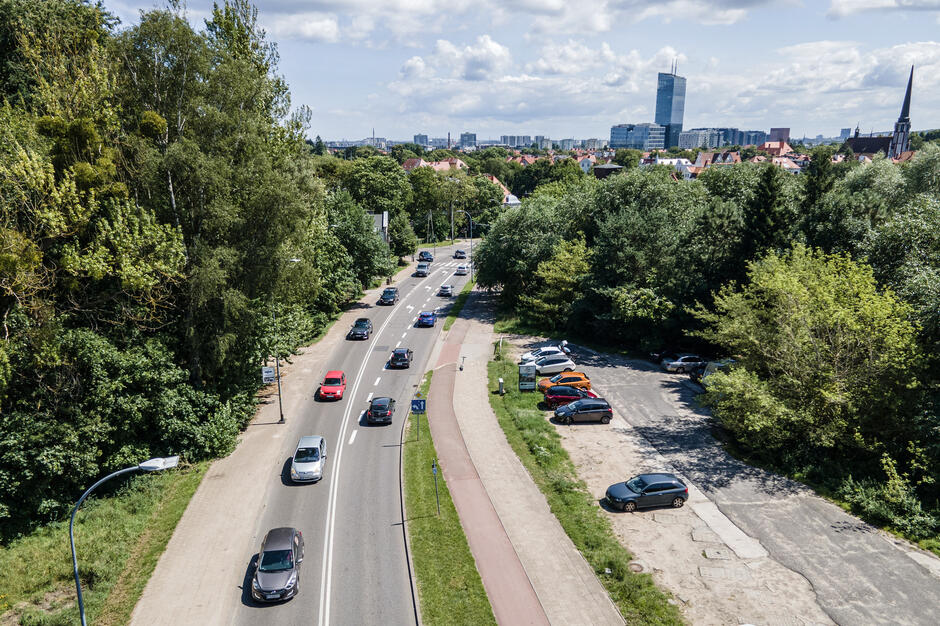 Zdjęcie przedstawia ruchliwą ulicę przebiegającą przez zieloną okolicę, z jednej strony otoczoną drzewami, a z drugiej poboczem z zaparkowanymi samochodami. W tle widać panoramę miasta z wysokimi budynkami i kościołem, co tworzy kontrast między naturą a zabudową miejską.