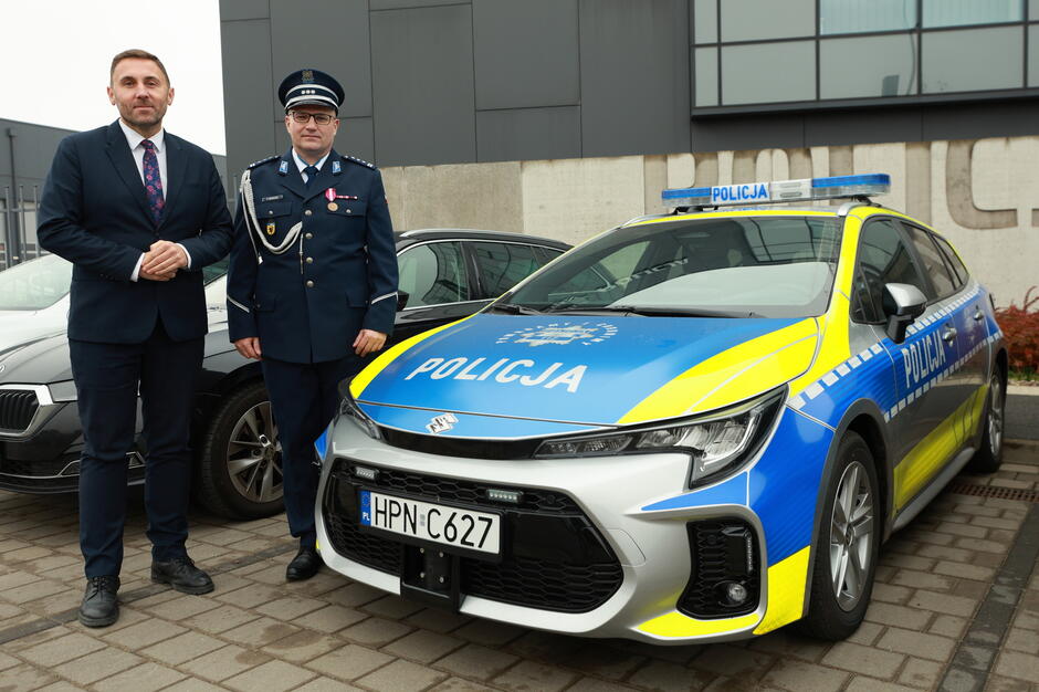 na zdjęciu oznakowany radiowóz, obok niego stoi policjant i mężczyzna w garniturze