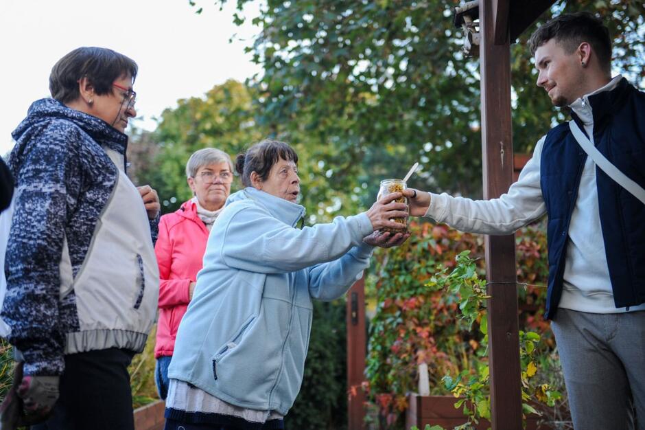 Starsze kobiety po lewej, jedna z nich przekazuje słoik młodemu mężczyźnie (po prawej)