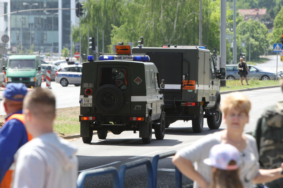 Na zdjęciu widać pojazdy patrolu saperskiego, zabezpieczające teren w mieście, prawdopodobnie w związku z wykryciem niewybuchu, co sugeruje napis Uwaga Niewybuchy  na samochodach. Wokół znajdują się cywile i służby, którzy zabezpieczają obszar, aby zapewnić bezpieczeństwo w razie zagrożenia.