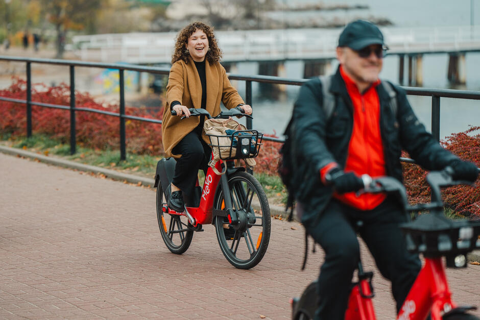 Na zdjęciu widać dwoje ludzi jadących na rowerach miejskich Mevo po alejce w parku. Kobieta z przodu, ubrana w brązowy płaszcz, śmieje się radośnie, podczas gdy mężczyzna w czapce i czerwonej bluzie jedzie obok niej na drugim planie.