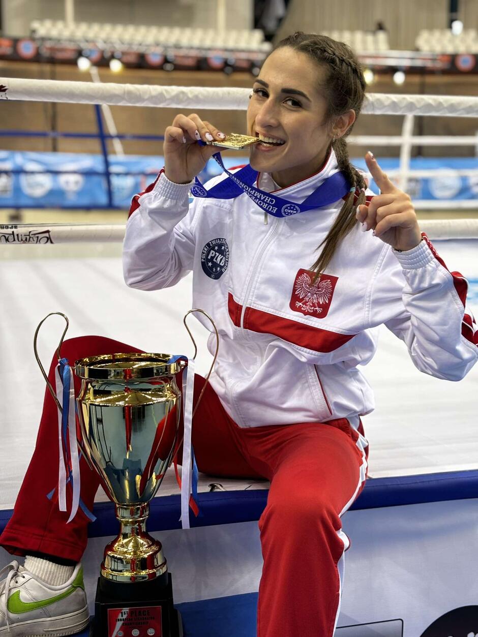 Kobieta w biało-czerwonym desie siedzi na krześle, w dłoni trzyma medal, który przygryza zębami. Obok stoi puchar