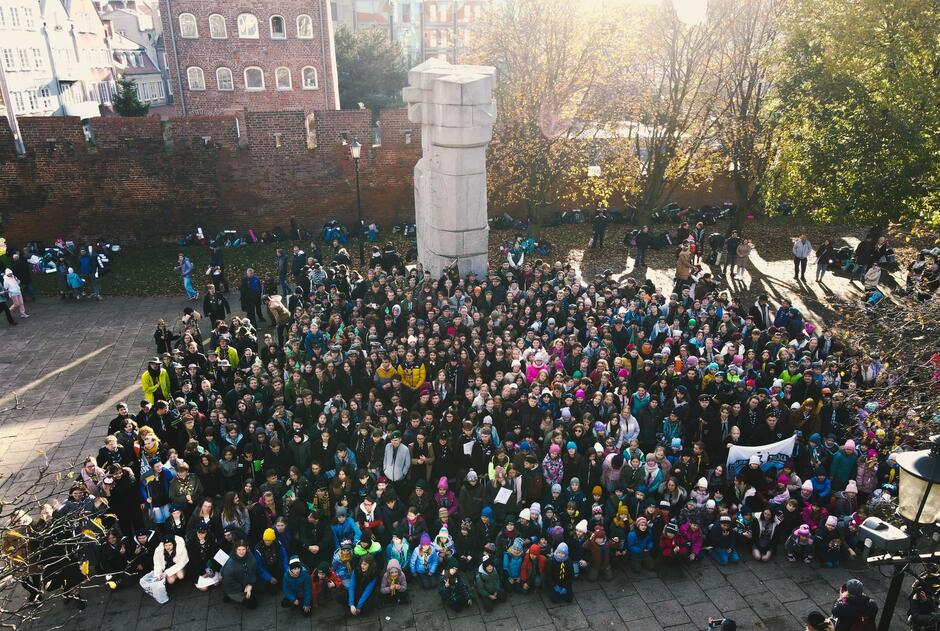 Duża grupa ludzi (ponad sto osób) stoi pod pomnikiem, pozują do zdjęcia