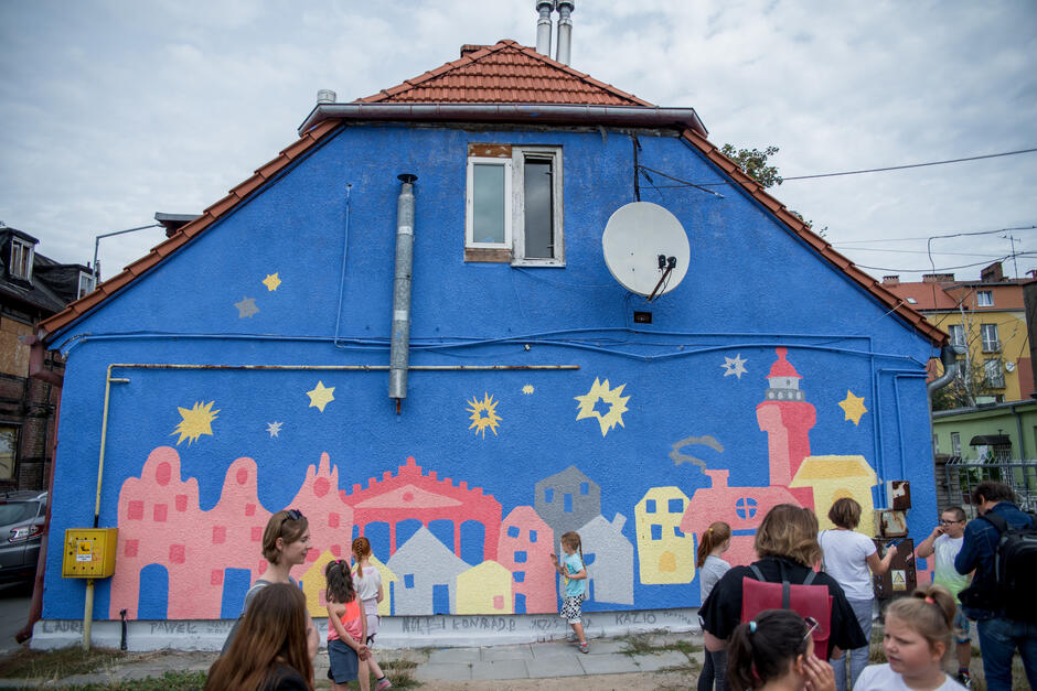 Niebiesko różowy obraz na fasadzie domu, przedstawia kolorowe budynki, na pierwszym planie dzieci oglądające mural - obraz.