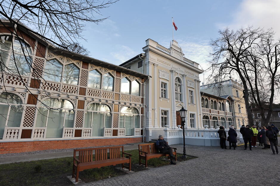 Na zdjęciu widoczny jest elegancki, klasyczny budynek o jasnej elewacji z flagą na dachu, otoczony zielenią i drzewami. Przed budynkiem stoi grupa ludzi spacerujących lub zwiedzających, co tworzy atmosferę spokojnego, miejskiego parku lub placu w historycznym otoczeniu.