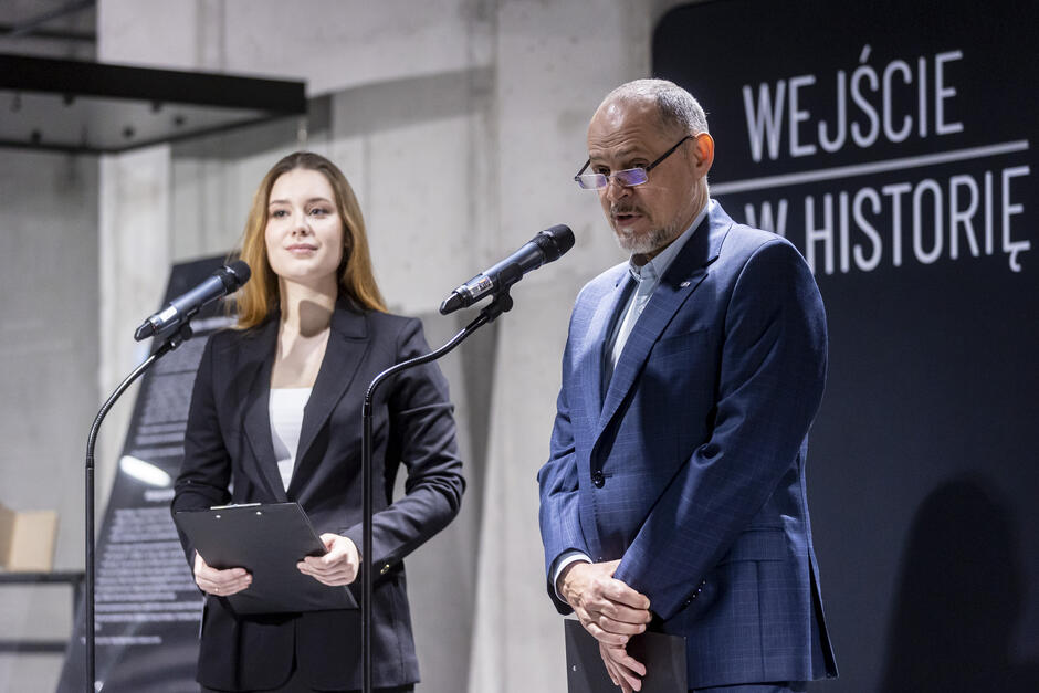 Na zdjęciu widać dwoje osób – kobietę i mężczyznę – stojących przy mikrofonach podczas oficjalnego wydarzenia. Oboje są elegancko ubrani; kobieta ma na sobie ciemny garnitur i trzyma w rękach notatki, a mężczyzna ubrany jest w niebieską marynarkę i również trzyma notatki. W tle znajduje się czarne tło z napisem „WEJŚCIE W HISTORIĘ”, co sugeruje, że jest to wydarzenie o charakterze historycznym lub związane z prezentacją wystawy. Obie osoby wyglądają profesjonalnie i skupione, co nadaje wydarzeniu poważny i oficjalny ton.