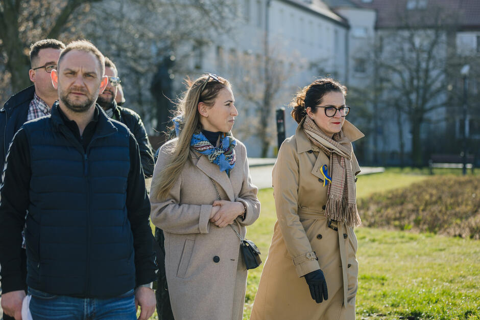 Na zdjęciu grupa ludzi ubranych w kurtki i płaszcze, na pierwszym planie dwie kobiety i mężczyzna, w tle zielony skwer