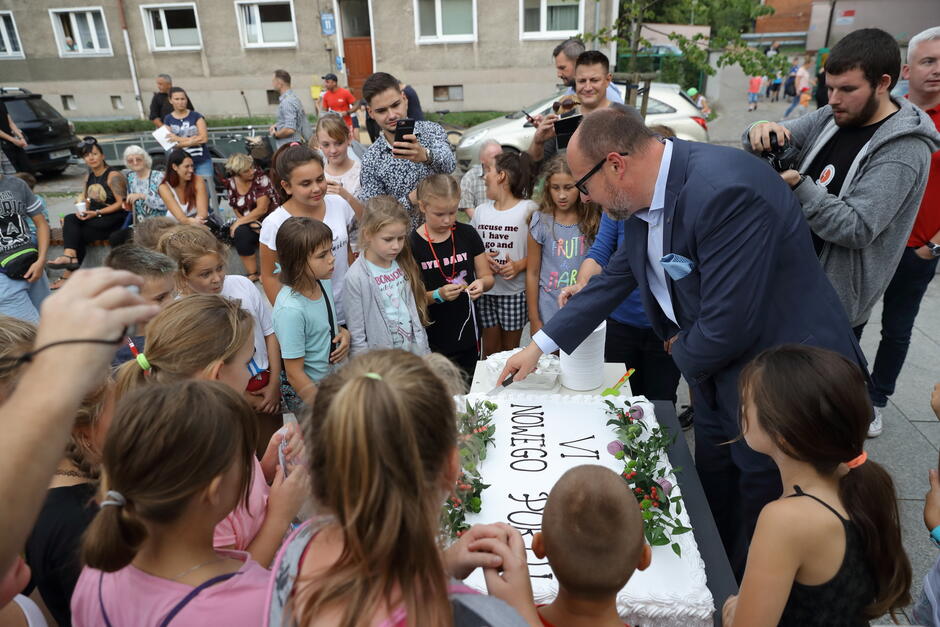 mężczyzna kroi tort, obok grupka dzieci, dzieci są ubrane kolorowo, uśmiechają się