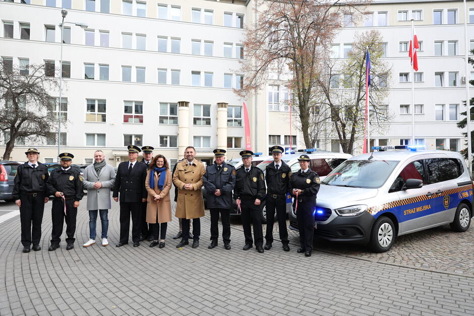 na zdjęciu stoi w jednym rzędzie ponad dziesięć osób, część z nich to strażnicy miejscy w mundurach, a także prezydent gdańska i jego zastępca, w tle wysoki budynek urzędu o jasnej elewacji