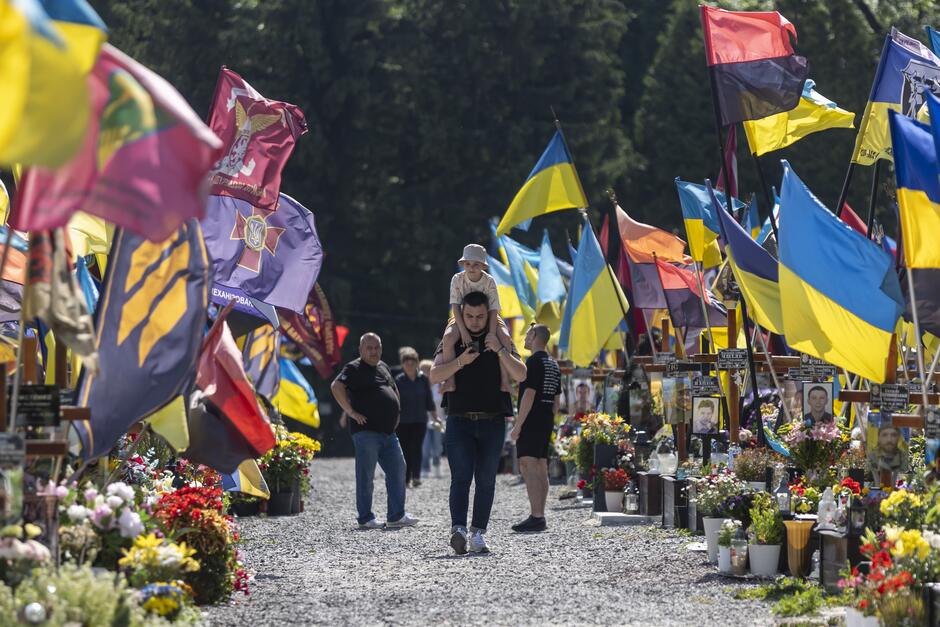 Aleja cmentarna, którą idą ludzie. Po lewej i prawej są groby żołnierzy i powiewające nad nimi niebiesko-żółte flagi