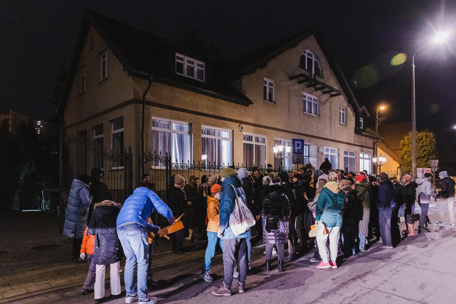 Na zdjęciu widać grupę ludzi zebranych przed dużym, jasnym budynkiem w nocy, oświetlonym przez uliczne latarnie