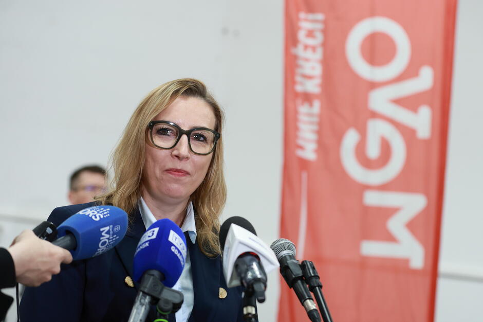 Na zdjęciu widoczna jest kobieta w okularach, przemawiająca do mikrofonów na konferencji prasowej. W tle znajduje się czerwona flaga z napisem MEVO oraz rozmazany sprzęt i osoby w tle, co sugeruje oficjalne wydarzenie.