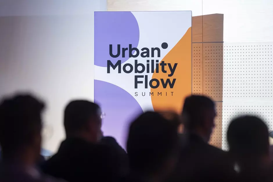 Na zdjęciu widnieje plansza z napisem Urban Mobility Flow Summit  na tle w odcieniach fioletu, bieli i pomarańczu, co sugeruje tematy związane z miejską mobilnością. Na pierwszym planie zarysowane są sylwetki uczestników, wskazując na wydarzenie lub konferencję dotyczącą rozwiązań mobilnych w miastach.