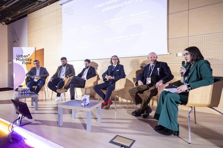 Na zdjęciu widoczna jest panelowa dyskusja podczas Urban Mobility Flow Summit, w której udział bierze sześć osób siedzących na scenie w formalnych strojach. Moderatorka po prawej stronie trzyma mikrofon, a za uczestnikami wyświetlany jest ekran z tytułem panelu oraz nazwiskami uczestników, co wskazuje na profesjonalną debatę dotyczącą zrównoważonego transportu miejskiego.