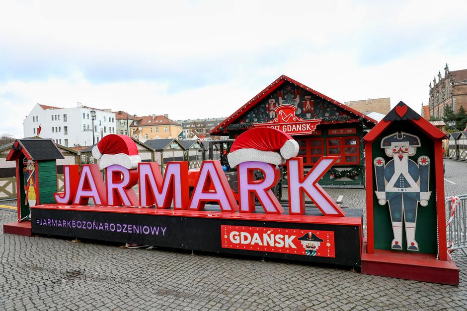 Zdjęcie przedstawia dekorację świątecznego Jarmarku Bożonarodzeniowego w Gdańsku, z dużym napisem „JARMARK” ozdobionym czapkami Mikołaja. W tle znajduje się świąteczny domek z ozdobami i grafikami, a po bokach widoczne są elementy dekoracyjne, takie jak figurka Dziadka do Orzechów