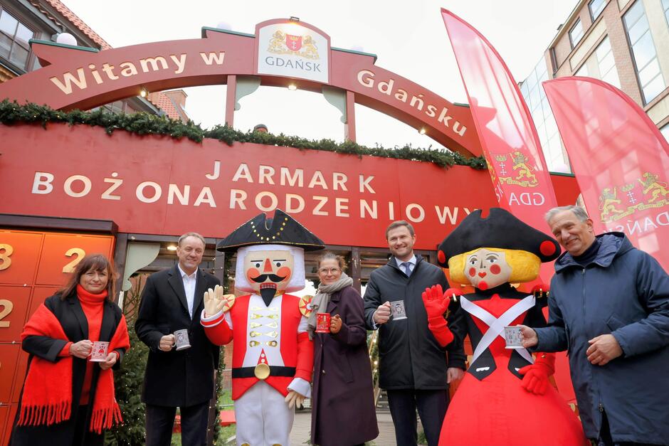 Zdjęcie przedstawia grupę osób stojących przed wejściem na Jarmark Bożonarodzeniowy w Gdańsku, ozdobionym czerwoną bramą z napisem „Witamy w Gdańsku”. Towarzyszą im postacie Dziadka do Orzechów i świątecznej lalki, a uczestnicy trzymają ozdobne kubki z motywami świątecznymi