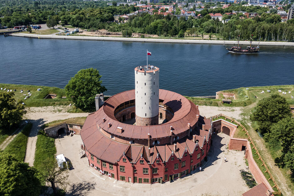 Na zdjęciu widoczna jest Twierdza Wisłoujście, historyczna budowla w Gdańsku, sfotografowana z lotu ptaka. Forteca otoczona jest wodami Motławy, a w tle widać fragment miasta oraz płynący po rzece statek stylizowany na jednostkę historyczną, co podkreśla turystyczny i zabytkowy charakter miejsca.