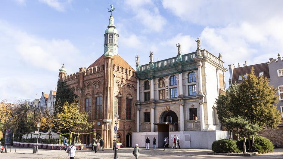 Na zdjęciu znajduje się fragment zabytkowej zabudowy Gdańska, przedstawiający dwa charakterystyczne budynki. Po lewej stronie widoczna jest Dwór Bractwa św. Jerzego, ceglany budynek w stylu gotyckim z charakterystycznymi łukami, ozdobnymi oknami i wieżyczką zwieńczoną kopułą oraz chorągiewką w kształcie smoka. Po prawej stronie widać Złotą Bramę, renesansowy budynek z jasną elewacją, kolumnami oraz bogato zdobionym gzymsem. Na balustradzie znajdują się rzeźby przedstawiające alegoryczne postacie.  Na pierwszym planie widoczni są spacerujący ludzie, a także drzewa i ogródki restauracyjne ozdobione roślinnością. Całość sceny rozgrywa się w pogodny dzień z niebieskim niebem i rozproszonymi chmurami.