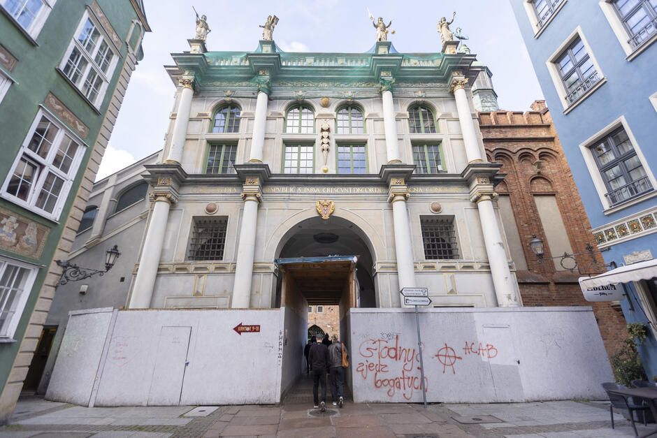 Na zdjęciu widoczna jest Złota Brama w Gdańsku, jeden z najbardziej charakterystycznych zabytków miasta, obecnie w trakcie remontu. Budynek w stylu manierystycznym wyróżnia się kolumnami, bogatymi zdobieniami i rzeźbami alegorycznymi na szczycie. Nad łukowym przejściem widnieje herb Gdańska oraz łacińska inskrypcja: Concordia res publicae parvae crescunt discordia magnae concidunt , co można przetłumaczyć jako:  Zgodą małe republiki rosną, niezgodą wielkie upadają.  Na pierwszym planie widać ogrodzenie remontowe z białej płyty, które zasłania dolną część bramy. Na ogrodzeniu znajdują się graffiti, w tym napisy takie jak:  Gdańsk zjedz bogatych  oraz różne symbole i tagi. Na środku ogrodzenia znajduje się prowizoryczne przejście, z którego korzystają przechodnie. Po bokach kadru widać fasady sąsiadujących kamienic – po lewej stronie zieloną, a po prawej niebieską. Całość fotografii utrzymana jest w perspektywie centralnej, co podkreśla symetrię zabytkowej bramy.