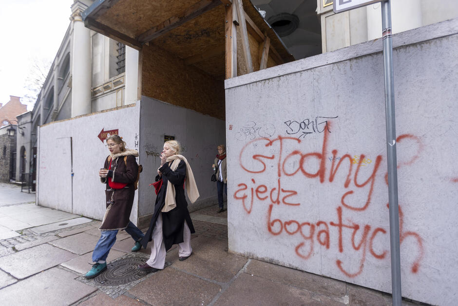 Na zdjęciu widoczna jest Złota Brama w Gdańsku, jeden z najbardziej charakterystycznych zabytków miasta, obecnie w trakcie remontu. Budynek w stylu manierystycznym wyróżnia się kolumnami, bogatymi zdobieniami i rzeźbami alegorycznymi na szczycie. Nad łukowym przejściem widnieje herb Gdańska oraz łacińska inskrypcja: Concordia res publicae parvae crescunt discordia magnae concidunt , co można przetłumaczyć jako:  Zgodą małe republiki rosną, niezgodą wielkie upadają.   Na pierwszym planie widać ogrodzenie remontowe z białej płyty, które zasłania dolną część bramy. Na ogrodzeniu znajdują się graffiti, w tym napisy takie jak:  Gdańsk zjedz bogatych  oraz różne symbole i tagi. Na środku ogrodzenia znajduje się prowizoryczne przejście, z którego korzystają przechodnie.  Po bokach kadru widać fasady sąsiadujących kamienic – po lewej stronie zieloną, a po prawej niebieską. Całość fotografii utrzymana jest w perspektywie centralnej, co podkreśla symetrię zabytkowej bramy.