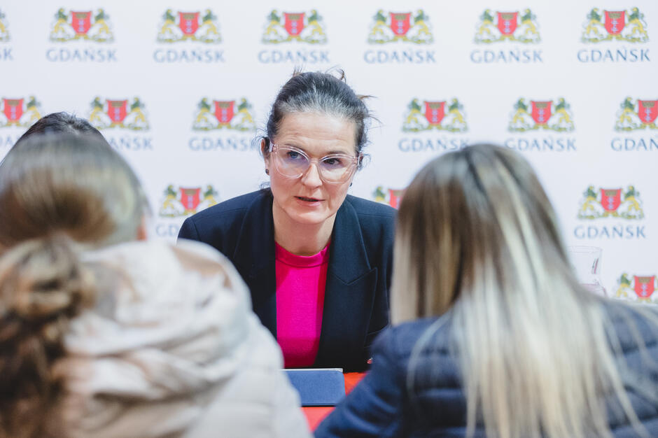 na zdjęciu siedząca prezydent gdańska w granatowej marynarce, widać ją od pasa w górę, naprzeciwko niej siedzą dwie kobiety w długich włosach, widać tyły ich głów