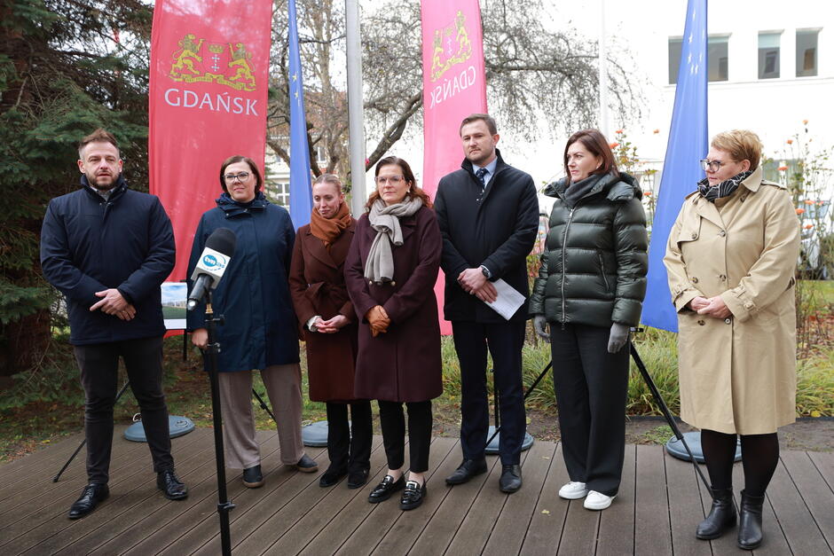 Na zdjęciu widoczna jest grupa osób stojących podczas konferencji prasowej, na tle flag miasta Gdańska oraz Unii Europejskiej. Osoby prezentują różnorodny strój formalny i casualowy, a za nimi znajduje się tablica z wizualizacją projektu budowlanego.