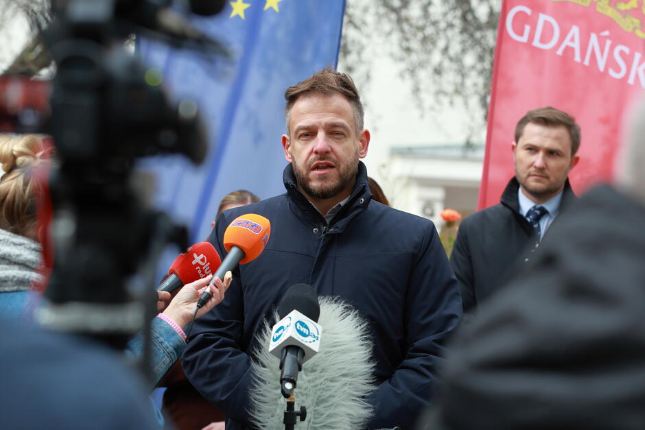 Na zdjęciu widać mężczyznę wypowiadającego się na konferencji prasowej, otoczonego mikrofonami różnych mediów. W tle widoczne są flagi, w tym flaga Gdańska, co sugeruje oficjalne wydarzenie miejskie.