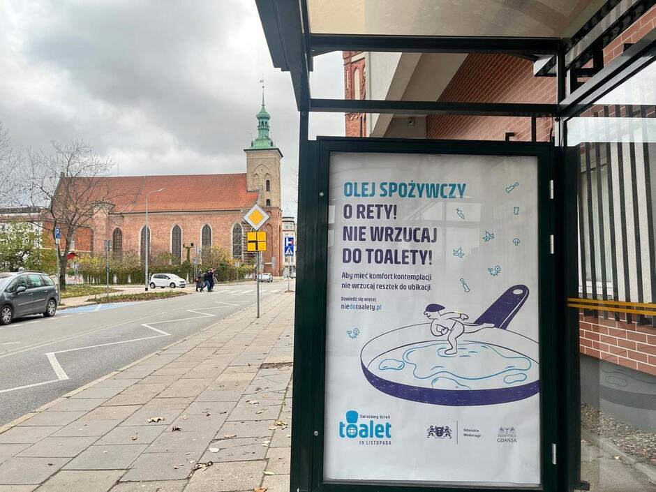 Na zdjęciu widzimy plakat zamieszczony na wiacie przystankowej, który apeluje o niewylewanie oleju spożywczego do toalety, aby zapobiec zanieczyszczeniom i awariom kanalizacji. W tle znajduje się ulica z zaparkowanymi samochodami oraz charakterystyczny budynek kościoła z wieżą.