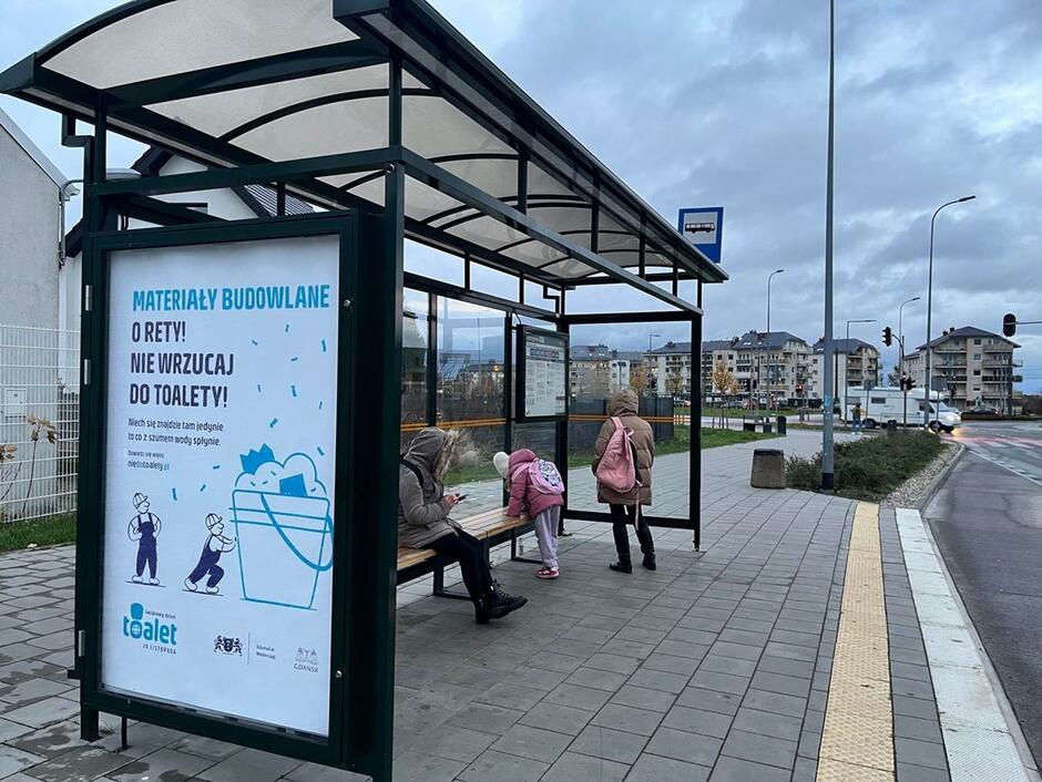 Na zdjęciu widzimy przystanek autobusowy z plakatem informacyjnym, który przestrzega przed wrzucaniem materiałów budowlanych do toalety, promując odpowiedzialne korzystanie z kanalizacji. W tle znajduje się osiedle mieszkaniowe oraz droga, a na przystanku siedzi kilka osób, chroniąc się przed chłodną pogodą.