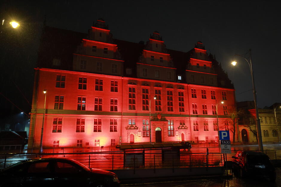 Na zdjęciu widzimy okazały budynek oświetlony intensywnym czerwonym światłem, co nadaje mu dramatycznego i symbolicznego charakteru. Sceneria nocna, deszczowa, z odbiciami świateł na mokrej nawierzchni, podkreśla wyjątkowy nastrój miejsca i wydarzenia.