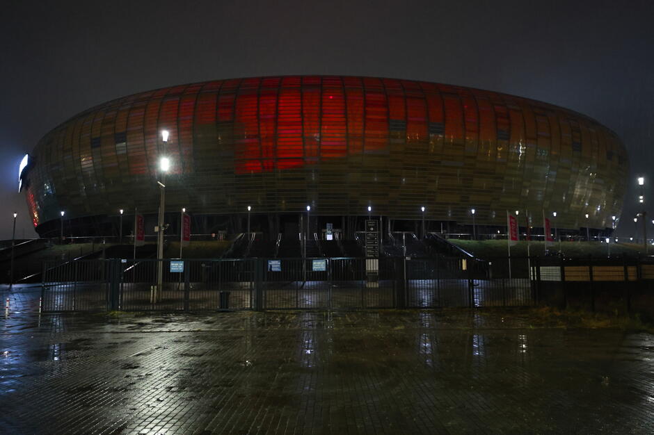 Na zdjęciu widoczny jest stadion podświetlony na czerwono, co tworzy efektowny i symboliczny wygląd. Otoczenie nocne i odbicia świateł na mokrej nawierzchni podkreślają atmosferę powagi i spokoju.