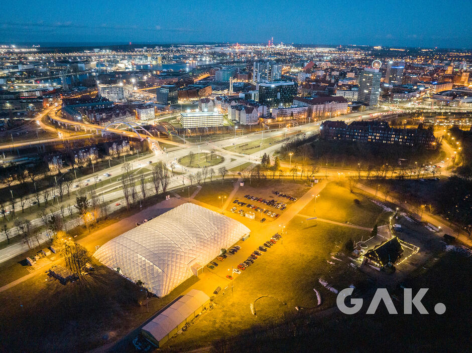 Zdjęcie przedstawia nocną panoramę miejską z widokiem na rozświetlone ulice, most oraz nowoczesne budynki, w tym obiekt o białej, kopułowej konstrukcji na pierwszym planie. Jasne światła miejskie oraz dobrze zorganizowana infrastruktura tworzą dynamiczny i nowoczesny obraz miasta.