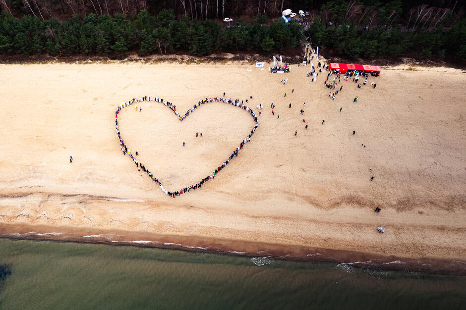 Na zdjęciu widoczna jest grupa ludzi ustawionych w kształt dużego serca na plaży, co symbolizuje solidarność i wspólnotę. W tle widać las i namioty organizatorów, a cała scena znajduje się w malowniczym nadmorskim krajobrazie.