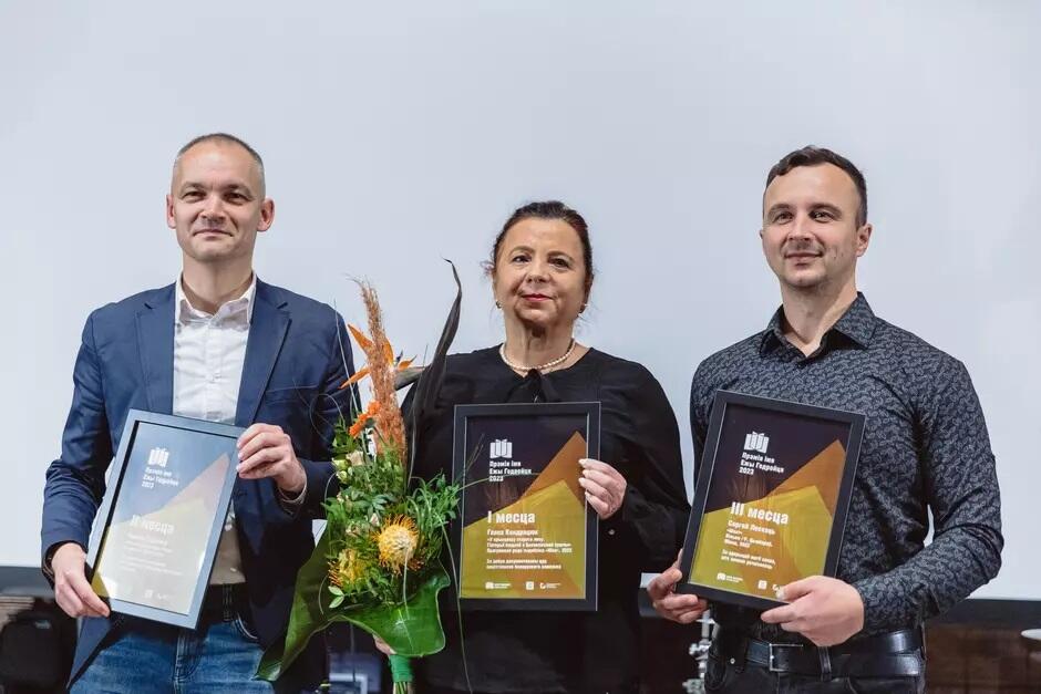 Na zdjęciu widzimy trzech laureatów poprzedniej edycji Niezależnej Nagrody Literackiej im. Jerzego Giedroycia, którzy dumnie trzymają w rękach swoje dyplomy. Kobieta w środku dodatkowo trzyma bukiet kwiatów, a wszyscy stoją na tle neutralnego, jasnego tła, uśmiechając się delikatnie