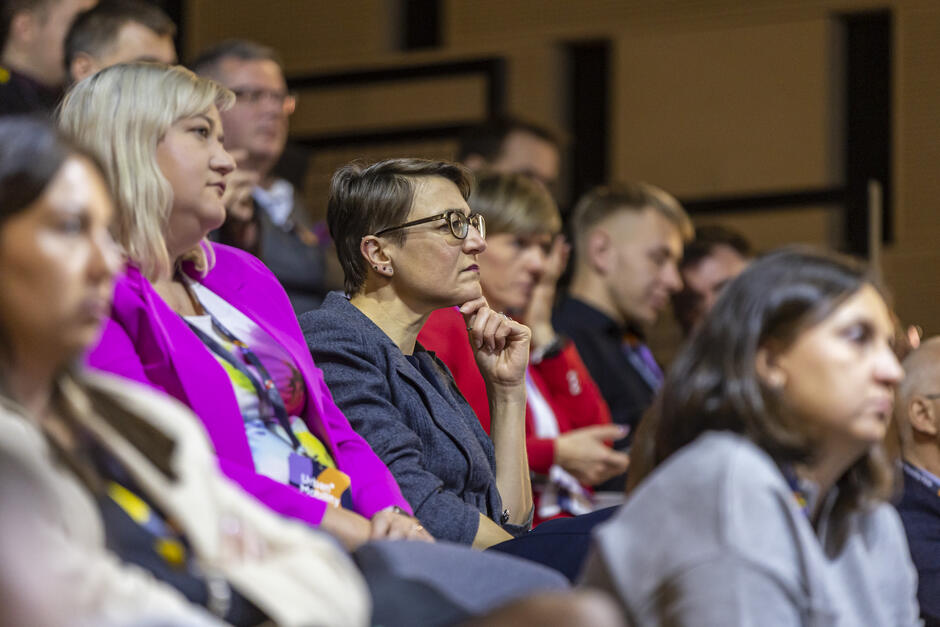 Na zdjęciu widać osoby siedzące w rzędach, uważnie uczestniczące w jakimś wydarzeniu, najprawdopodobniej konferencji lub prelekcji. W centrum uwagi znajduje się kobieta w okularach i szarym stroju, skupiona i zamyślona, opierająca rękę na brodzie.