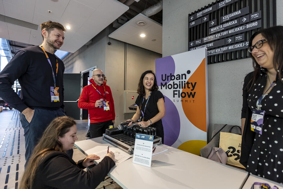 Na zdjęciu widoczna jest grupa osób przy stoisku oznaczonym logiem „Urban Mobility Flow Summit”, gdzie odbywa się rejestracja lub wypożyczanie sprzętu, np. słuchawek do tłumaczeń. Uczestnicy są w dobrych nastrojach, rozmawiają i śmieją się, co wskazuje na przyjazną i profesjonalną atmosferę wydarzenia.