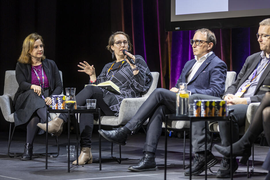 Zdjęcie przedstawia panel dyskusyjny, w którym uczestniczą cztery osoby siedzące na scenie w wygodnych fotelach. Jedna z uczestniczek trzyma mikrofon i gestykuluje, prowadząc rozmowę, podczas gdy pozostali paneliści słuchają z uwagą, a na stolikach przed nimi znajdują się szklanki z wodą i materiały promocyjne.